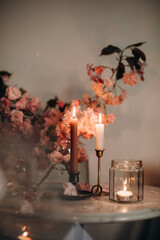 burning tall candles in vintage candlesticks. candlelight near a transparent vase with pink branches with flowers. home evening comfort and warmth.