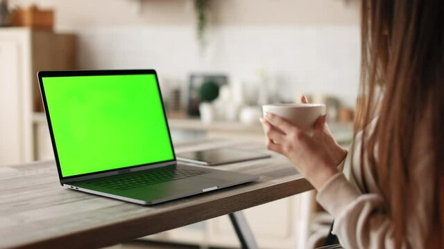 A woman is sitting in her home office. She is drinking tea. On the table is a laptop with a green screen. Chromakey. 4K