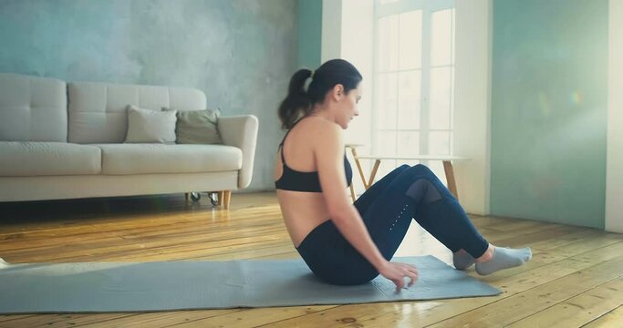 Young woman in stylish sportswear does crunches and leg lifts on grey mat in living room with parquet slow motion
