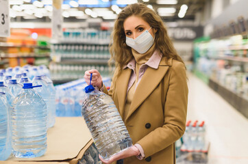 A young woman buys bottled water in a supermarket. Woman in facial mask and gloves to prevent...