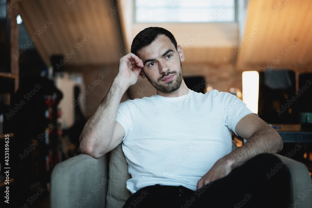 Wall mural portrait of handsome serious young brunette man in white t-shirt sitting in armchair at home