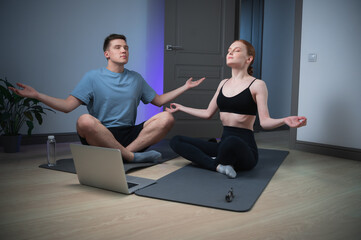 During an online yoga class, a young couple meditates on gym mats at home in front of a laptop