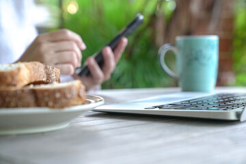 Person surfing on her phone with computer laptop on table. Working while having breakfast at home.