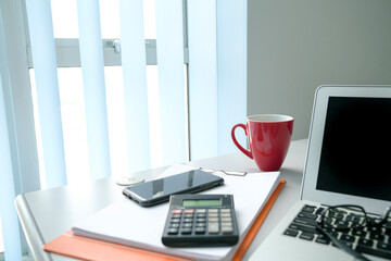 Office desk with computer laptop, phone, calculator, documents and cup of coffee. By the windows with vertical blinds. 