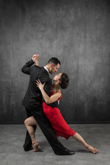 Couple of professional dancers in elegant suit and red dress in a tango dancing movement on dark background. Handsome man and woman dance looking  eye to eye. - obrazy, fototapety, plakaty