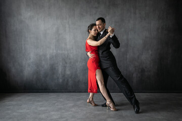 Couple of professional dancers in elegant suit and red dress in a tango dancing movement on dark background. Handsome man and woman dance cheek to cheek. - obrazy, fototapety, plakaty