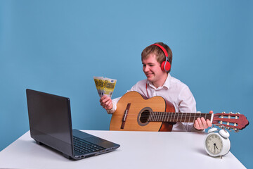 A man wearing headphones with a guitar and a computer on a blue background holds money in euros,...