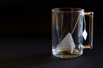 A white pyramid tea bag with green tea lies in a transparent heat-resistant glass mug on a black background. Free space for an inscription