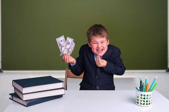 A Cunning Boy With Money In His Hands At A Desk In A School Classroom. US Dollars In Junior's Hand On A Blackboard Background, Copy Space For Text