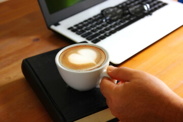 hand holding morning coffee in relax time . heart shape surface on white coffee cup on black book with  office supplies