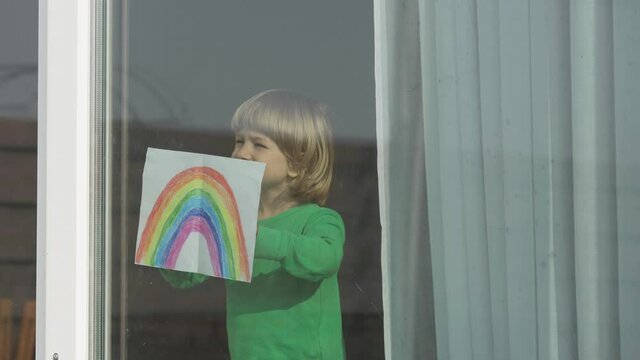 Blond hair little child with rainbow drawing at window, looking around for hope, conceptual