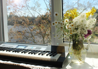 On the windowsill there is a synthesizer and a vase with white and yellow daisies and lilacs....