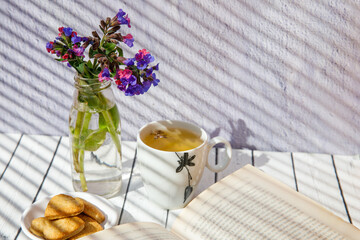 Romantic composition of bouquet pink and purple wild forest Lungwort, bisquits, cups green or herbal tea, old book on white wooden background with light shadows. Copy space for text, horizontal