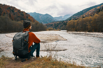 woman with a backpack in a jacket and a hat on the river bank in the mountains side view