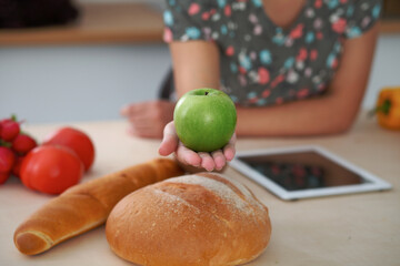 Close-up of female hand holding green apple in kitchen interiors. Many vegetables and other meal at glass table are ready for been cooked soon