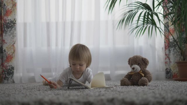 Portrait of little child drawing at notebook close to teddy bear toy lying together on carpet, home activity, isolation home
