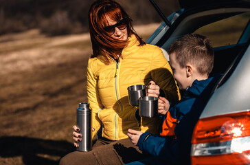 Mother and son take break from driving sitting by the car while drinking tea in nature.
