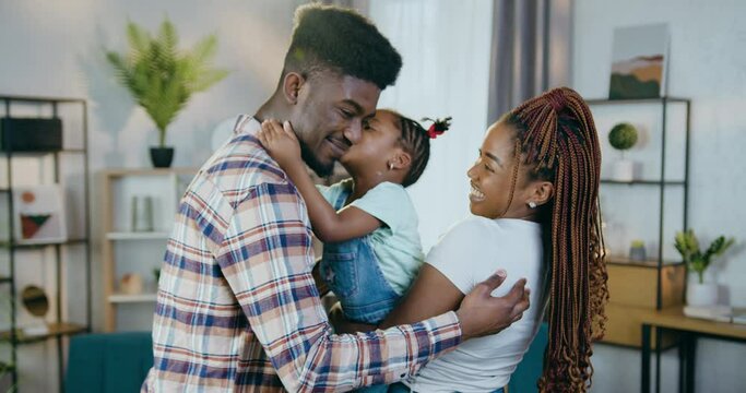 Young african parents kissing and gently embracing their cute little daughter. Positive family enjoying happy weekends together at home.