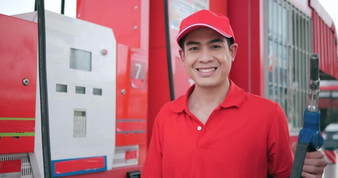 Portrait of Asian worker in red uniform with fuel pump nozzle looking camera at gas station service