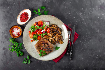 Grilled beef steak, well done. With mushrooms and fresh vegetable salad on a plate on the dark gray background top view