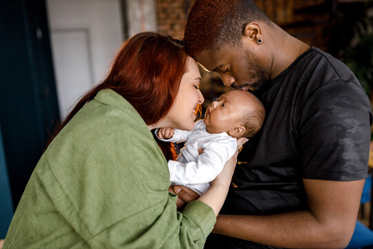 Happy Multiracial Mixed Family Gently Holding Newborn Baby. Red Haired Caucasian Mom Looking At Her Child, Afro American Father Lovingly Kissing Cute Baby In Head