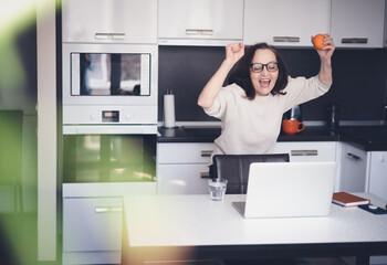 Beautiful happy mature elderly woman with orange in her hands standing at home in the kitchen in front of laptop screen, healthy eating and active retirees concept