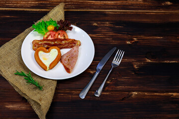 Closeup shot of delicious tasty American breakfast heart shape fried egg bacon ham fresh tomato with green salad vegetable on white plate decorated on sack placed with knife and fork on wooden table