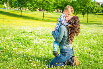 Fototapeta premium happy mom and child chilling in public park garden in summer. mother and son relaxing toghether outdoor sitting on grass playing. togetherness, love, family and happiness concept . sweet kiss portrait