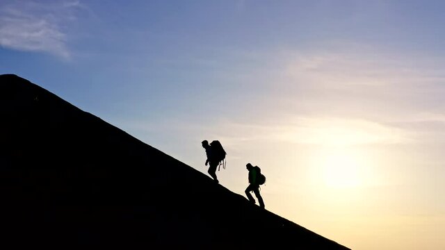 Silhouettes of a man and a woman with backpacks climbing to the top of the mountain.