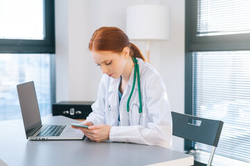 happy smiling female doctor in white coat using mobile phone sitting at desk with laptop in modern office of medic clinic on background of window in sunny day.