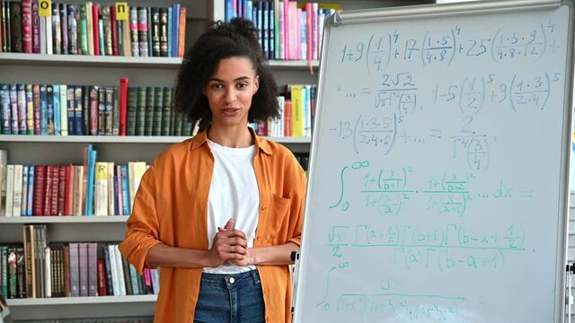 Modern African American smart young adult female teacher stand near the whiteboard, conducts online lecture by a video conference, looks at the webcam, shows information to the students, smiles