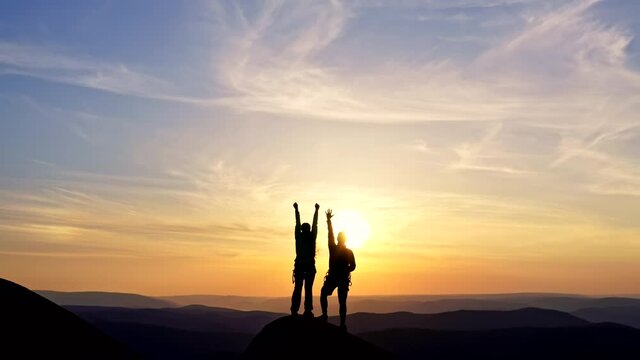 A young couple cheerfully raises their hands while standing on a mountaintop at sunset.