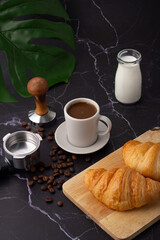 A white coffee cup was placed next to a milk bottle and a croissant on a cutting board, coffee beans and grinders on a marble floor.
