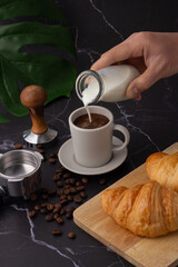The hand was holding a milk bottle pouring it into a white coffee cup, croissant on a cutting board, coffee beans and a coffee grinder on a black marble floor.