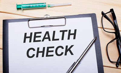 The words HEALTH CHECK is written on a white piece of paper next to black-rimmed glasses, a pen and a syringe. Medical concept