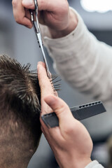 Professional barber making haircut to young man using scissors and comb at barbershop. Close-up, selective focus