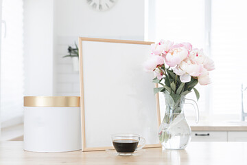 Fototapeta na wymiar Vertical frame and gift box mockup on a wooden table in the kitchen. Glass vase with a bouquet of pink peonies and a cup of black coffee. Scandinavian style interior.