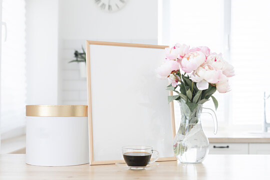 Vertical frame and gift box mockup on a wooden table in the kitchen. Glass vase with a bouquet of pink peonies and a cup of black coffee. Scandinavian style interior.