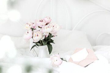 Obraz na płótnie Canvas Bouquet of pink peonies and greeting envelope on white bed linen. Modern interior in the bedroom. Wedding and festive style.