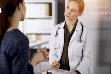 Cheerful smiling female doctor and patient woman discussing current health examination while sitting in sunny clinic. Perfect medical service in hospital. Medicine concept