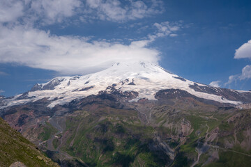 Fototapeta premium Volcano Elbrus, its peaks are slightly shrouded in clouds in sunny weather. Landscape view in the south-east of mount from Cheget mount. Kabardino-Balkaria region, Russia.