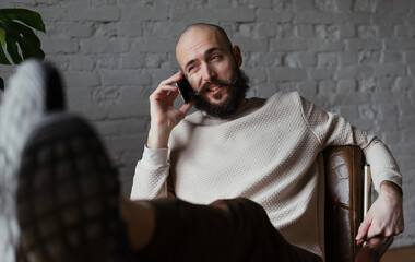 bald man 35 years old with a beard and mustache in a beige jumper sits and talks on the phone