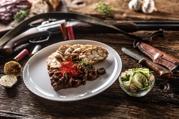 Porcelain plate full of deer venison goulash stew served with dumpling and fresh salad as an appetizer. Food is surrounded by hunting accesories such as hunting shotgun, bullets and rustic knife
