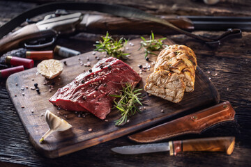 Nutrient rich raw deer venison prepared for a cooking process on a rustic wooden desk with roasted garlic, rosemary and huntig accesories like shot gun and ammunition - obrazy, fototapety, plakaty