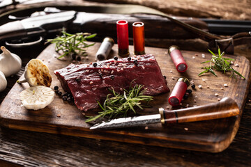 Nutrient rich raw deer venison prepared for a cooking process on a rustic wooden desk with roasted garlic and huntig accesories like shot gun and ammunition - obrazy, fototapety, plakaty