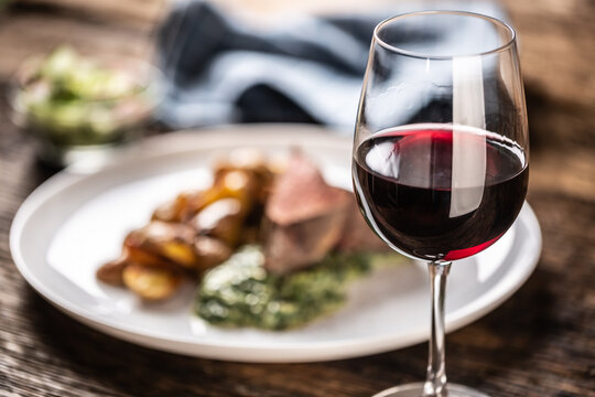 Red wine as a typical baverage to a red meat steak with roasted potatoes, spinach dip and fresh salad served on a white porcelain plate on the wooden desk.