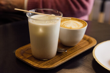 Iced Thai tea latte in glass mug and capuccino with wooden tray saucer on wooden table in a coffee shop.