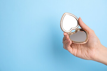 Woman holding stylish heart shaped cosmetic pocket mirror on light blue background, closeup. Space...