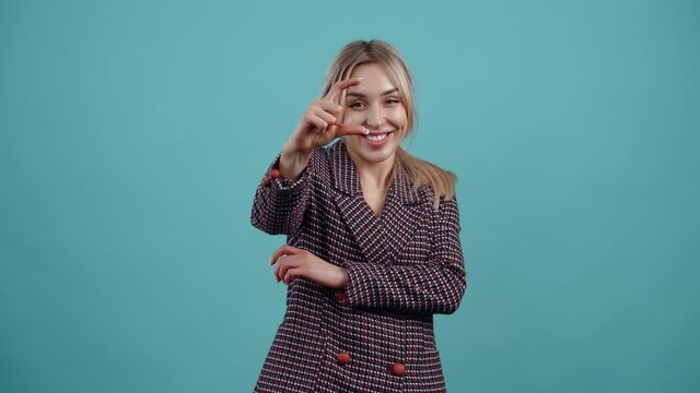 The blonde, amused by her blond hair, shows with her hand the gesture of a small size and displays small smiles. Isolated on a turquoise background. The concept of life. People's emotions. Portrait 4k
