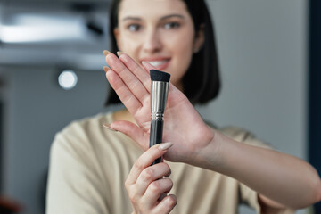Pretty young woman smiling and showing her favorite makeup brush in focus. Gorgeous brunette female making an online tutorial with beauty product. - obrazy, fototapety, plakaty